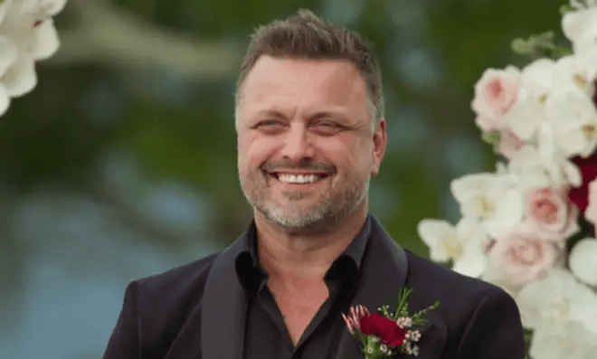 A smiling man in a tuxedo at an outdoor wedding.