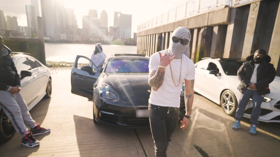 Screengrab of a music video showing a man in a balaclava and several other people standing near luxury cars.