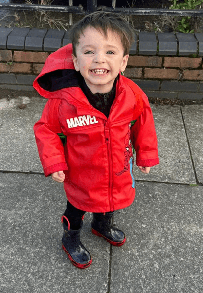 Smiling toddler in a red Marvel raincoat and rain boots.