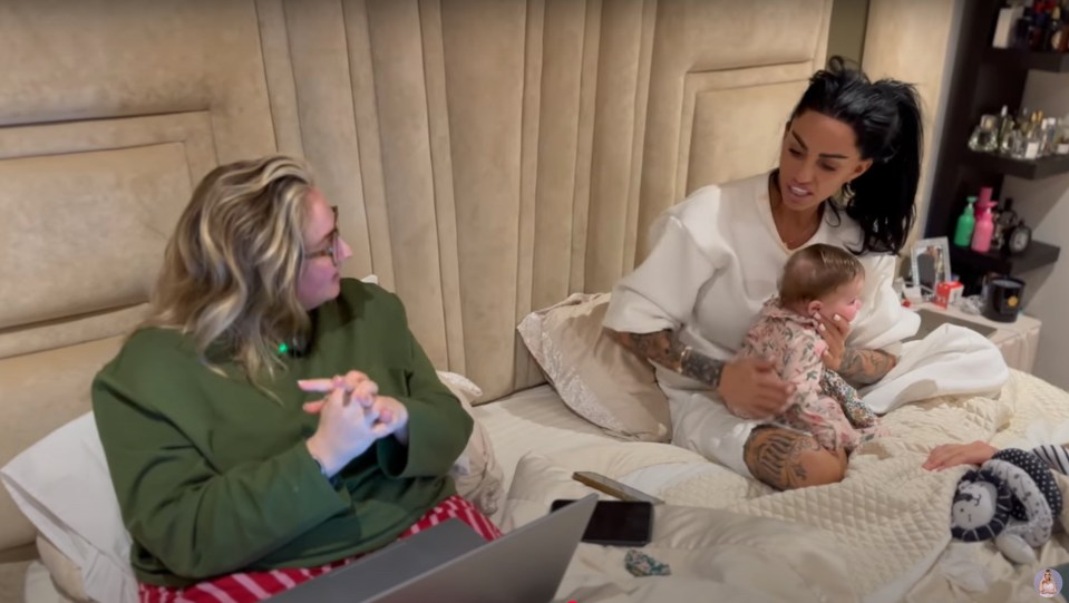 Katie Price sits on a bed with a baby, talking to a woman using a laptop.