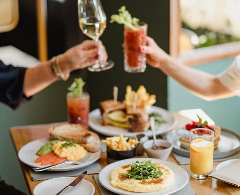 Two people toasting with drinks at a brunch table with various food items.