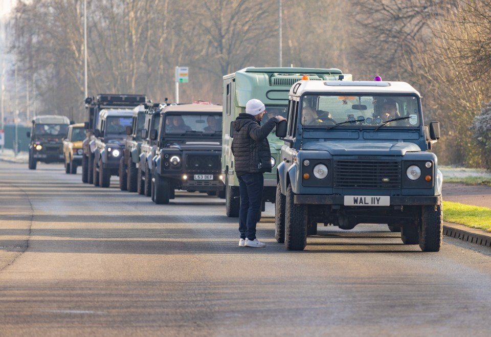 A line of Land Rovers driving down a road, with a person talking to the driver of one of them.