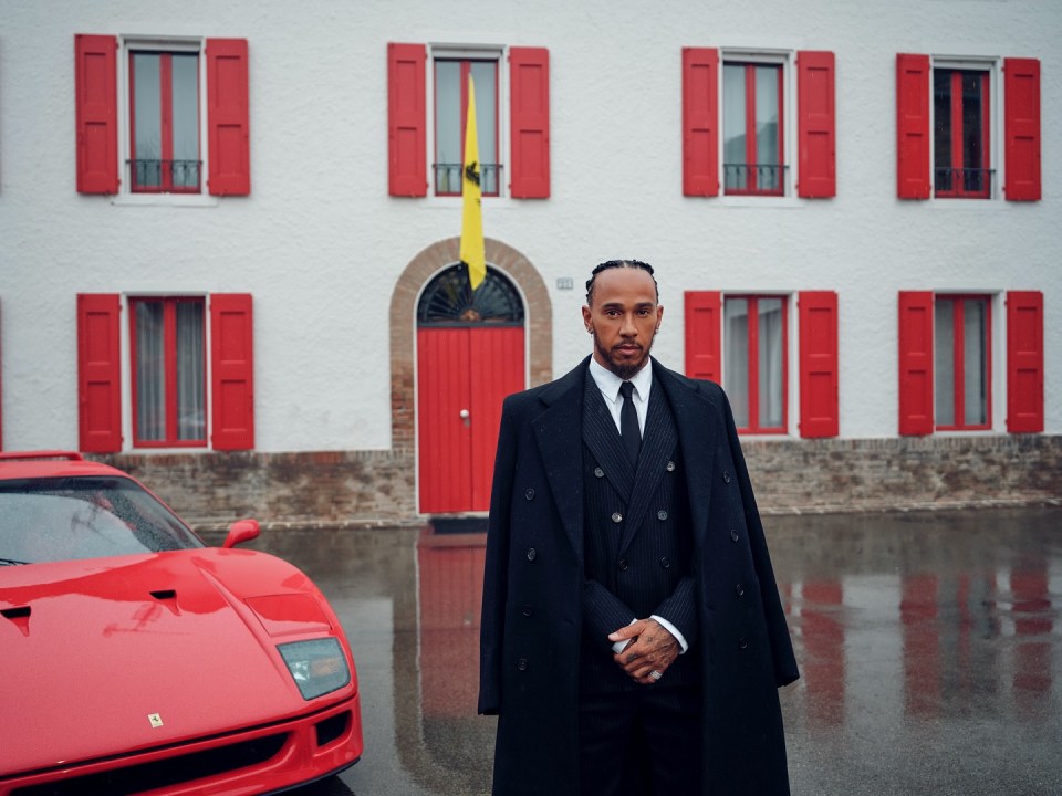 Lewis Hamilton in a black suit and overcoat, standing in front of a red Ferrari.