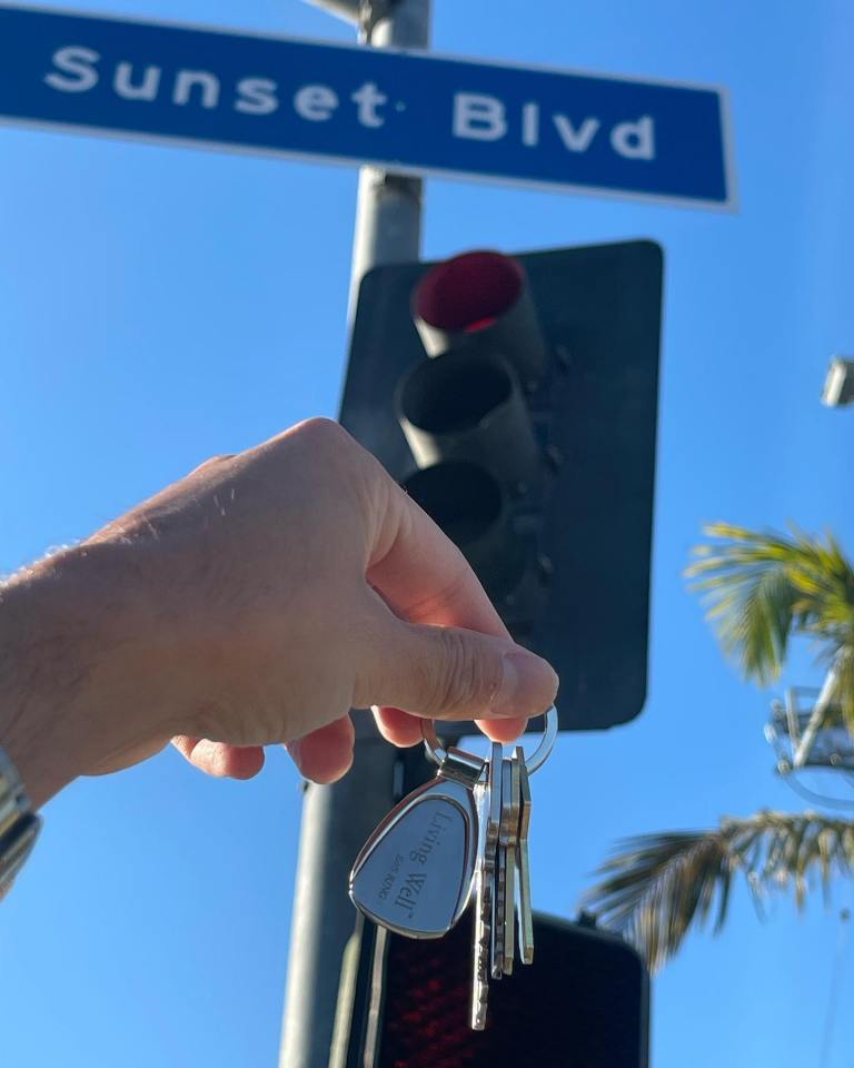 Hand holding keys with "Living Well" engraved on a keychain in front of a Sunset Blvd street sign.