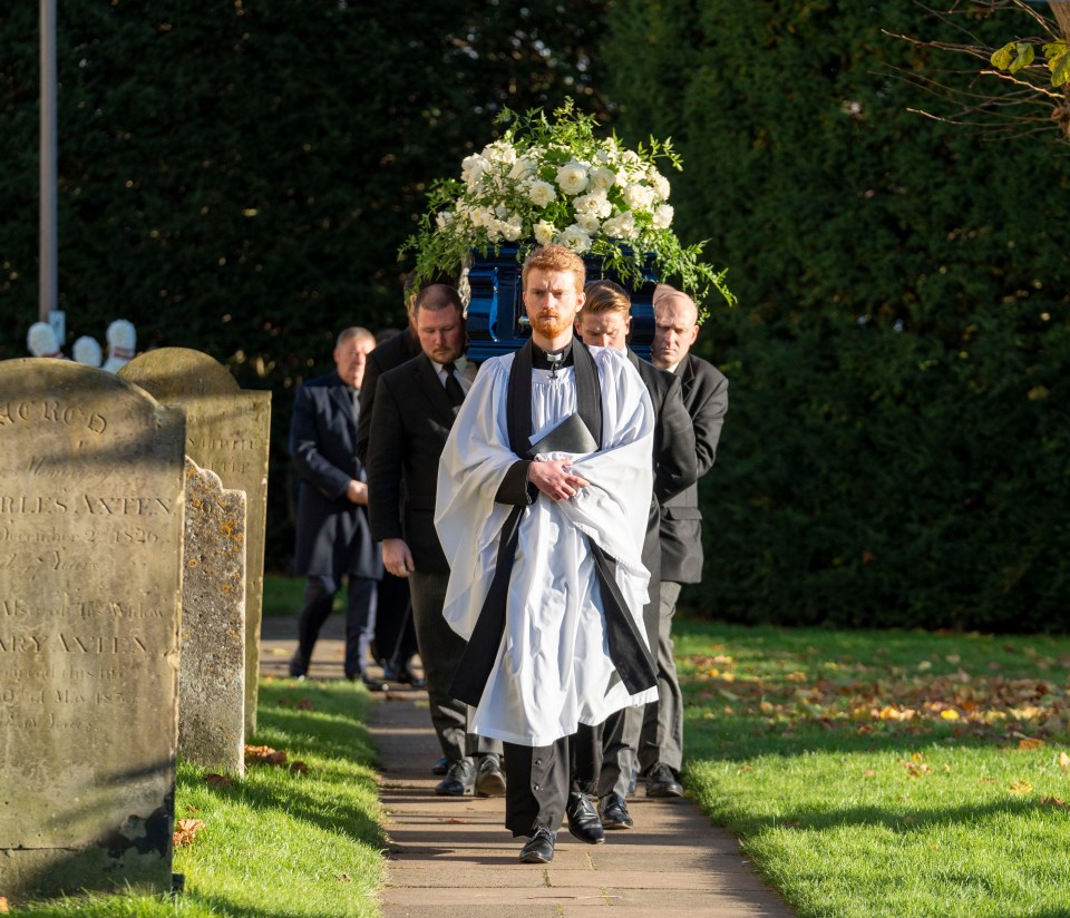 A coffin, adorned with white flowers, is carried by pallbearers during a funeral procession.