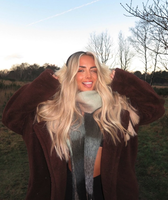 Abi Moore sticking her tongue out while wearing a brown coat and scarf.