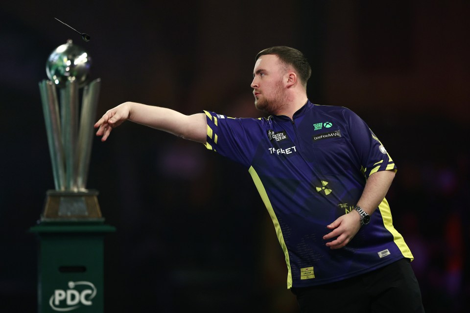 Luke Littler throwing a dart at a darts championship trophy.