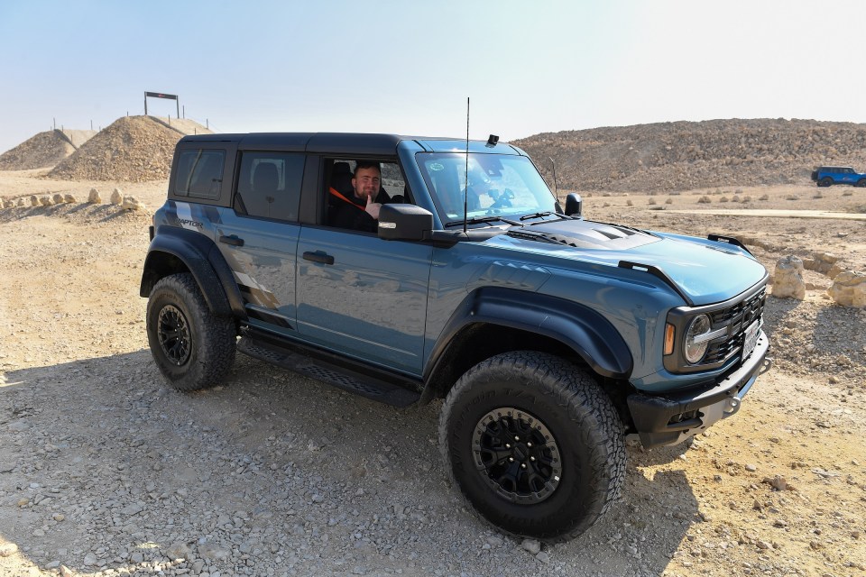 Man giving thumbs up while driving a blue Ford Bronco Raptor in the desert.