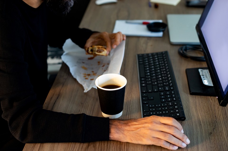 Man eating a pastry and drinking coffee at his office desk.