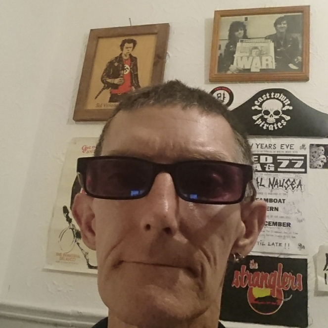 Close-up selfie of a person wearing sunglasses, with punk rock memorabilia on the wall behind them.