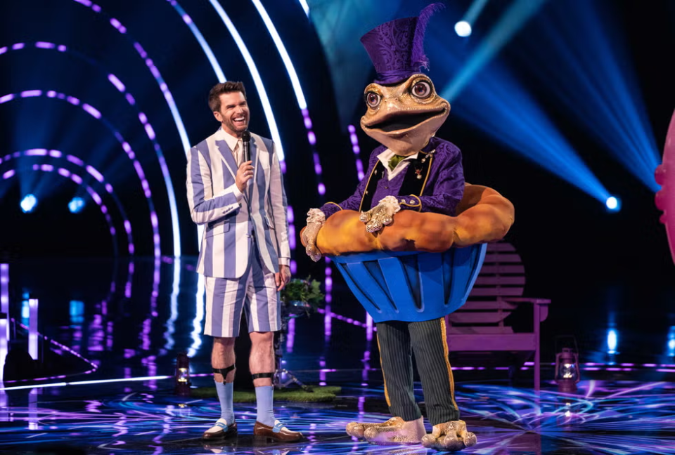 A man in a striped suit holds a microphone, standing next to a frog-like contestant in a cupcake costume on the Masked Singer stage.