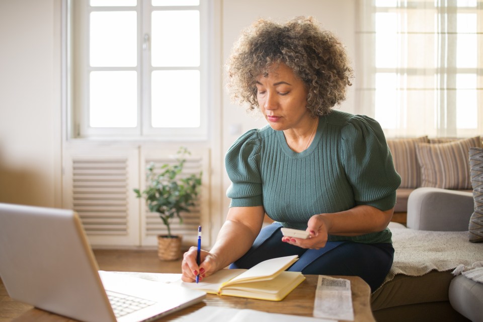 A middle-aged woman sits on a couch, paying bills using a laptop and calculator.