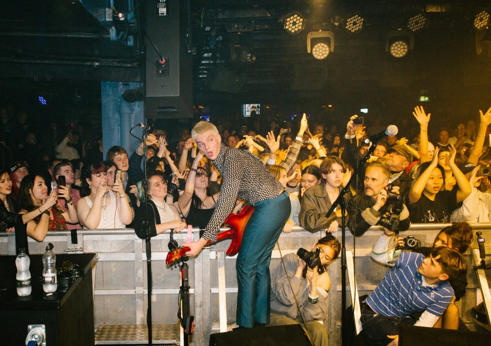 A musician with blond hair playing a red guitar on stage in front of a crowd.