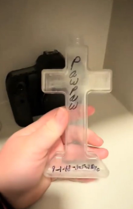 A hand holding a clear plastic cross with "Pawpaw" written on it.