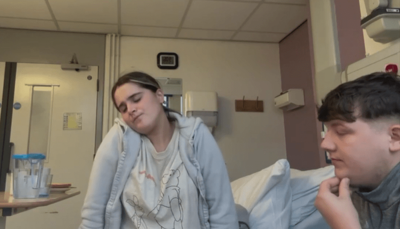 A young woman rests her head on her shoulder in a hospital room while a young man sits beside her.