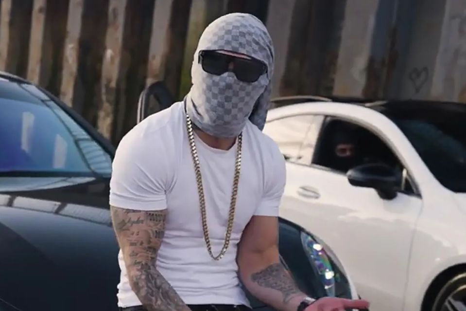 Man wearing a patterned balaclava and gold chain, sitting near two cars.