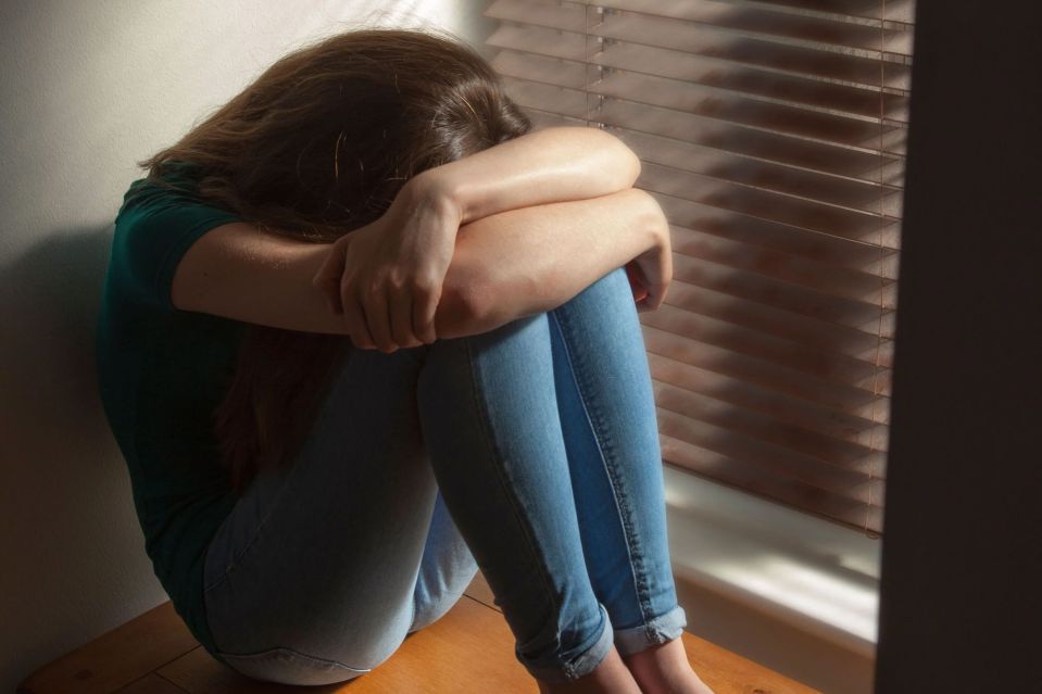 A young woman sits by a window, hugging her knees and hiding her face.