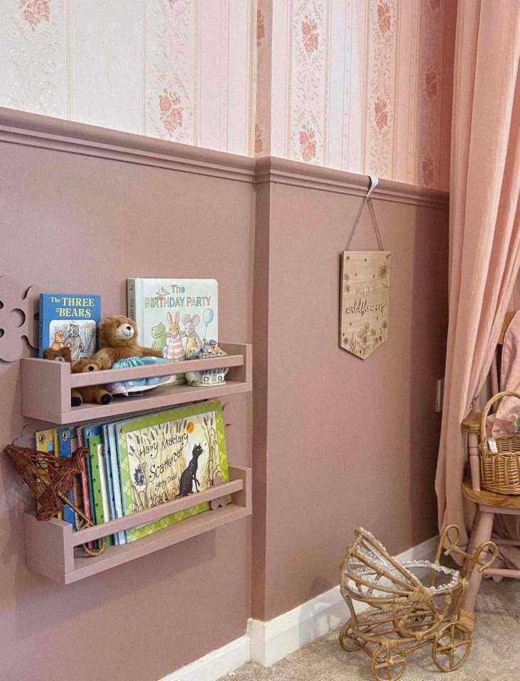 A girl's bedroom with pink walls, floral wallpaper, and a pink shelf holding children's books and toys.