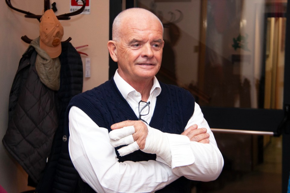 Portrait of a man with his arms crossed, wearing a white shirt and dark blue sweater vest; his right hand is bandaged.