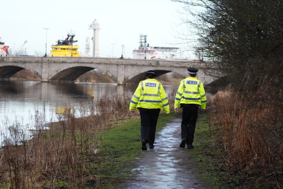 Two police officers walk along a path next to a river.