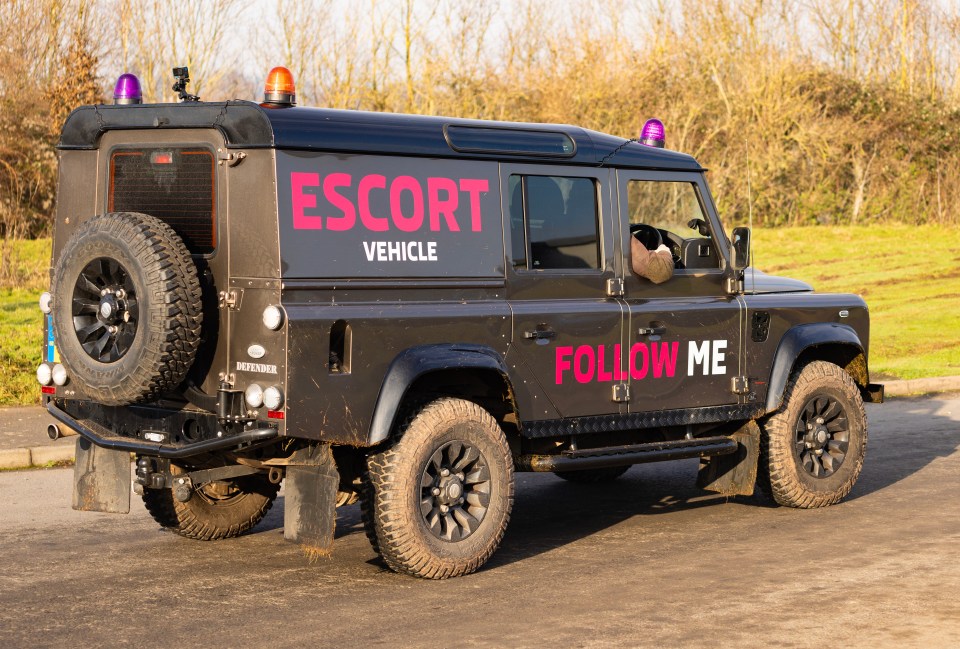 A dark-colored Land Rover Defender, labeled "Escort Vehicle" and "Follow Me," is driven by a person whose arm is visible in the window.