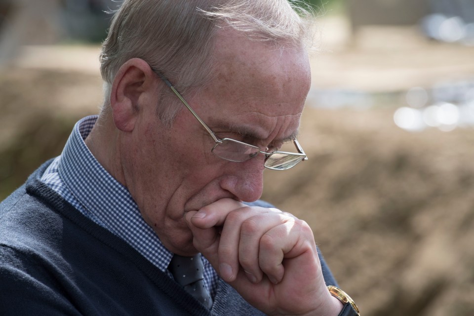 A man with glasses contemplates, hand to mouth.