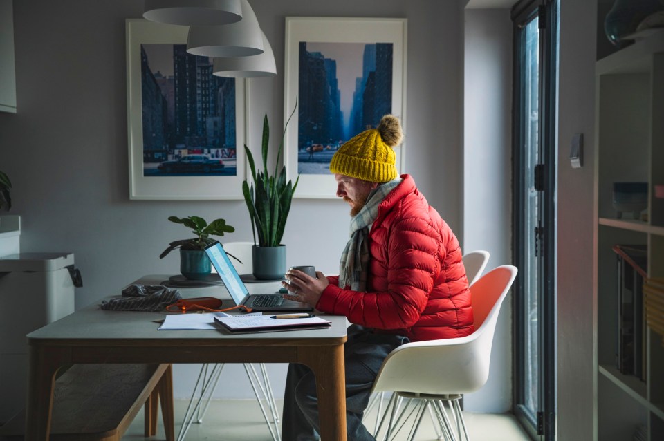 Man working on laptop at home wearing winter clothes to stay warm due to high energy costs.