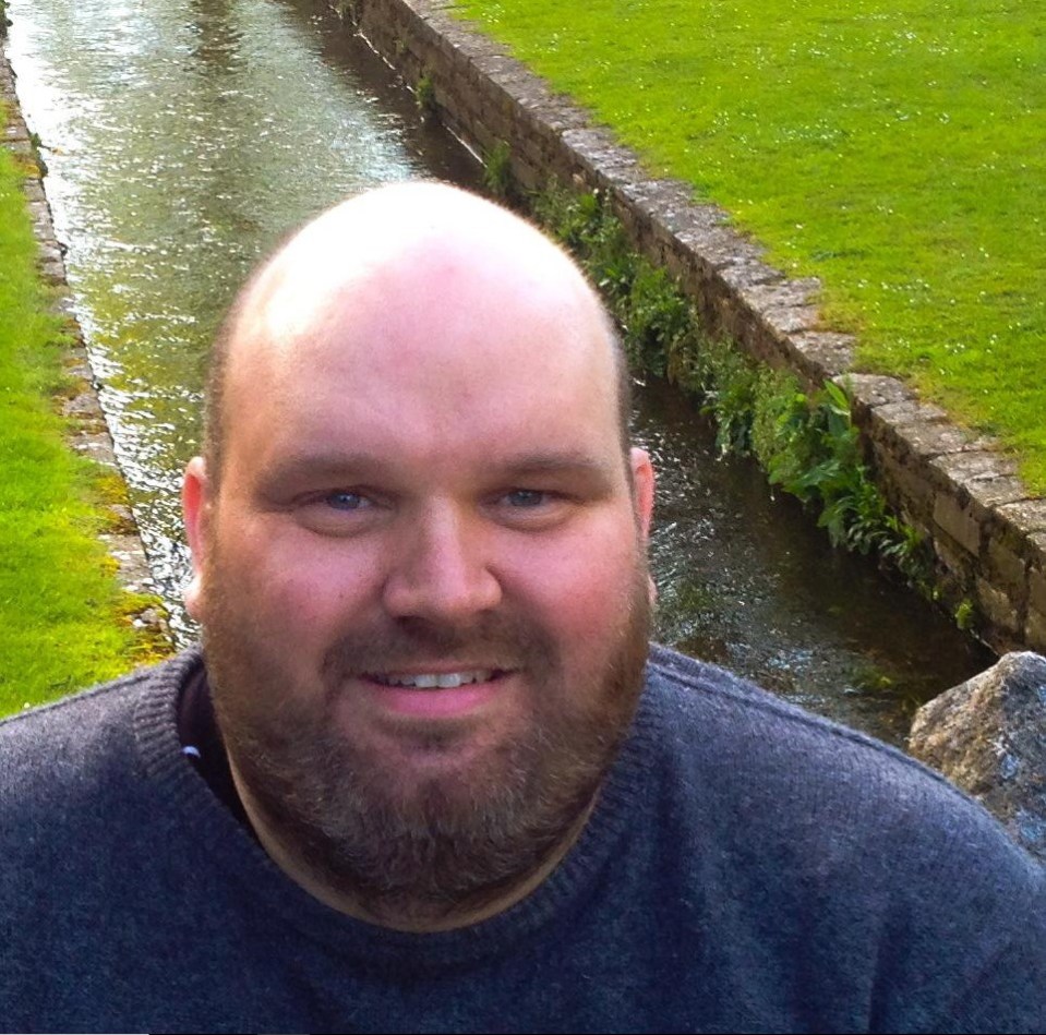 Photo of a man with a beard, smiling, near a stream.