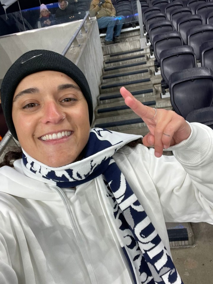 A woman wearing a white hoodie and a navy and white scarf makes a peace sign in a stadium.