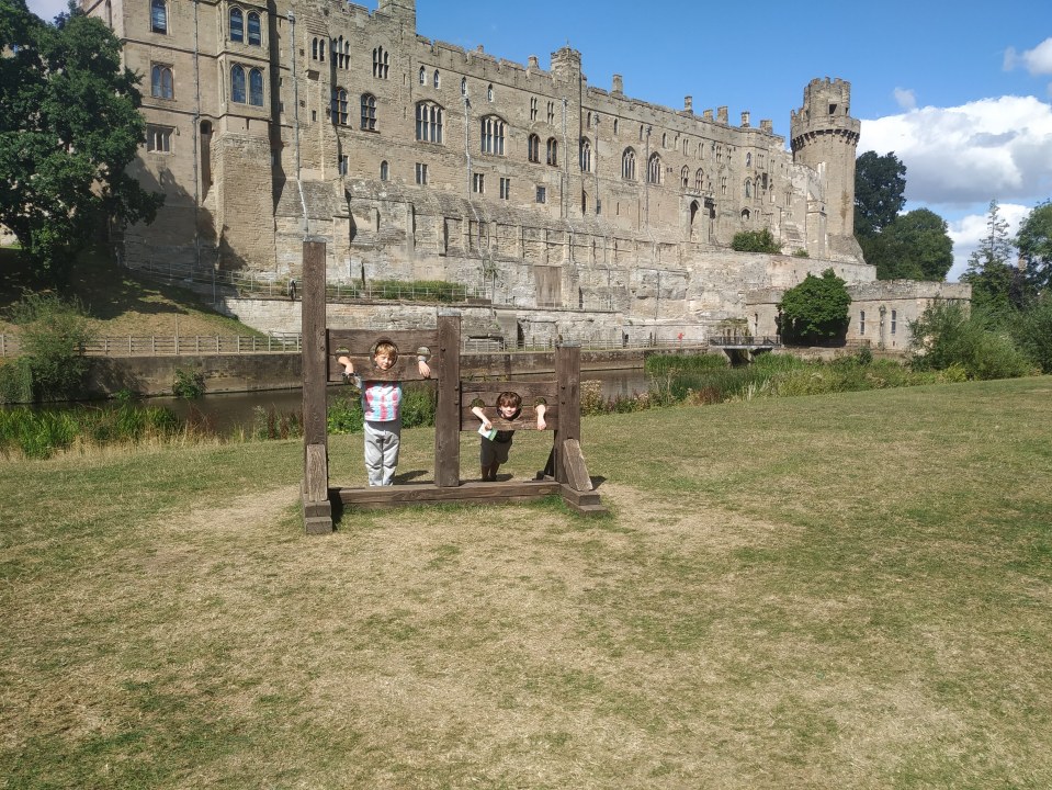 Two boys standing in a wooden pillory in front of a castle.