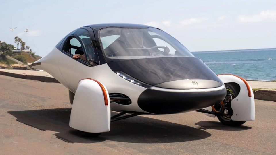 Three-wheeled solar-powered car driving near the ocean.