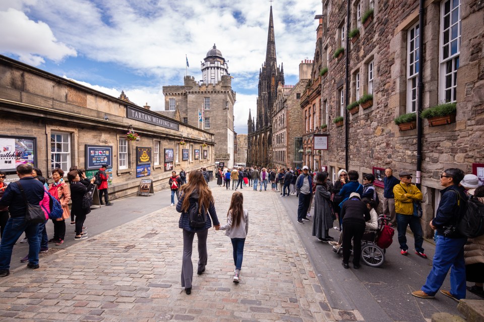 Tourists walking along a cobblestone street in Edinburgh, Scotland, past shops and historical buildings.