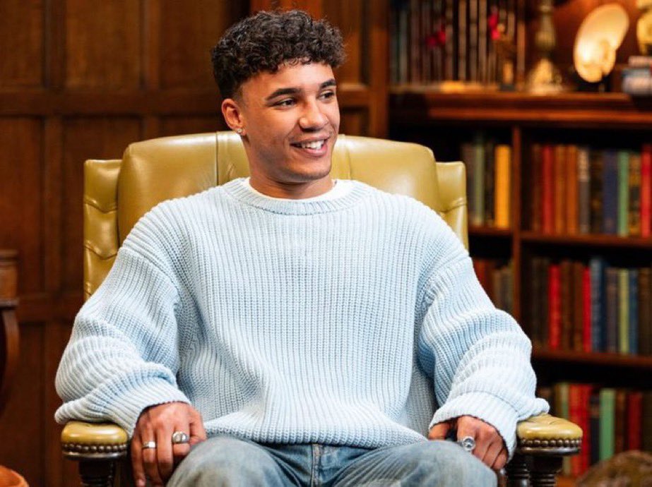 A young man in a light blue sweater sits in a chair.