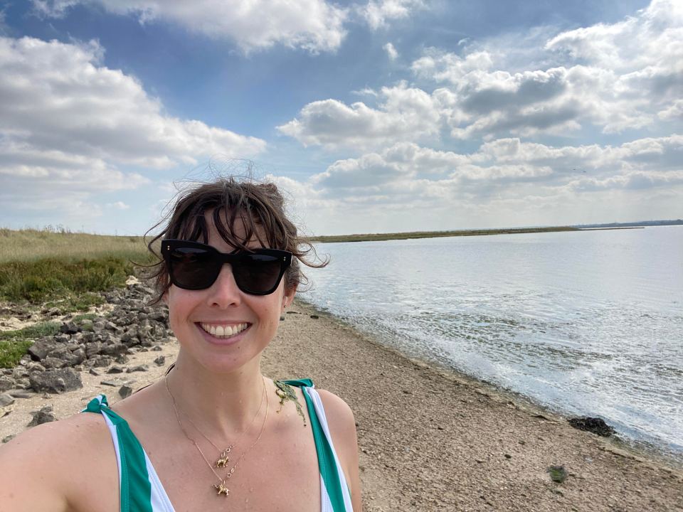 Woman in sunglasses smiling on a beach by the water.