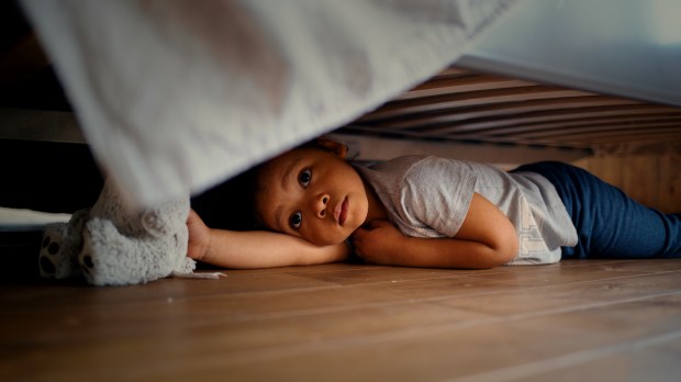 Toddler lying under a bed, holding a stuffed animal.