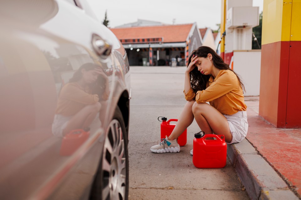 Woman sitting on the curb at a gas station, looking distressed, with two red gas cans beside her.