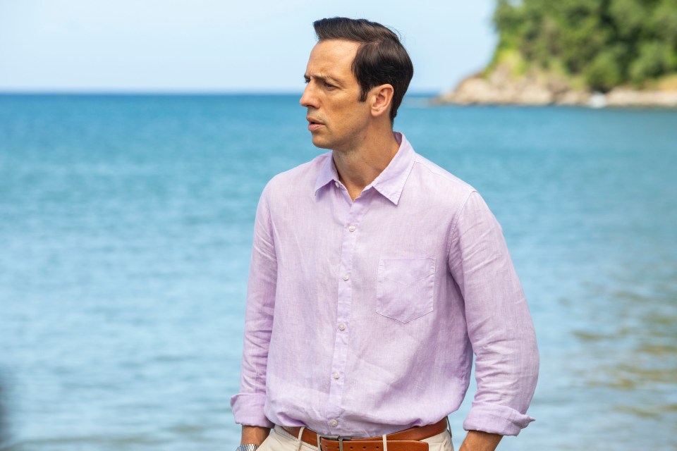 Ralf Little, portraying DI Neville Parker in Death In Paradise, standing by the ocean.