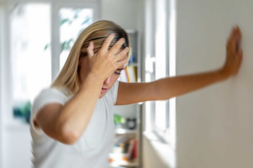 Woman experiencing vertigo, holding her head and leaning against a wall.