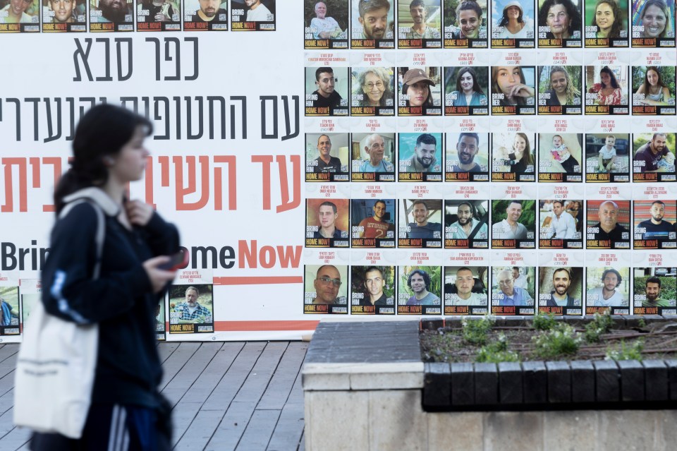 A woman walks past a wall displaying photos of hostages with the text "Bring them home now!" in Hebrew and English.