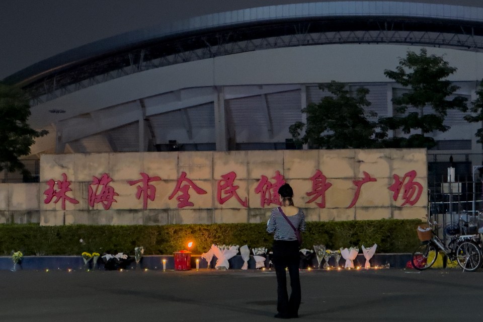 A woman stands before a memorial of flowers and candles in front of a stadium.