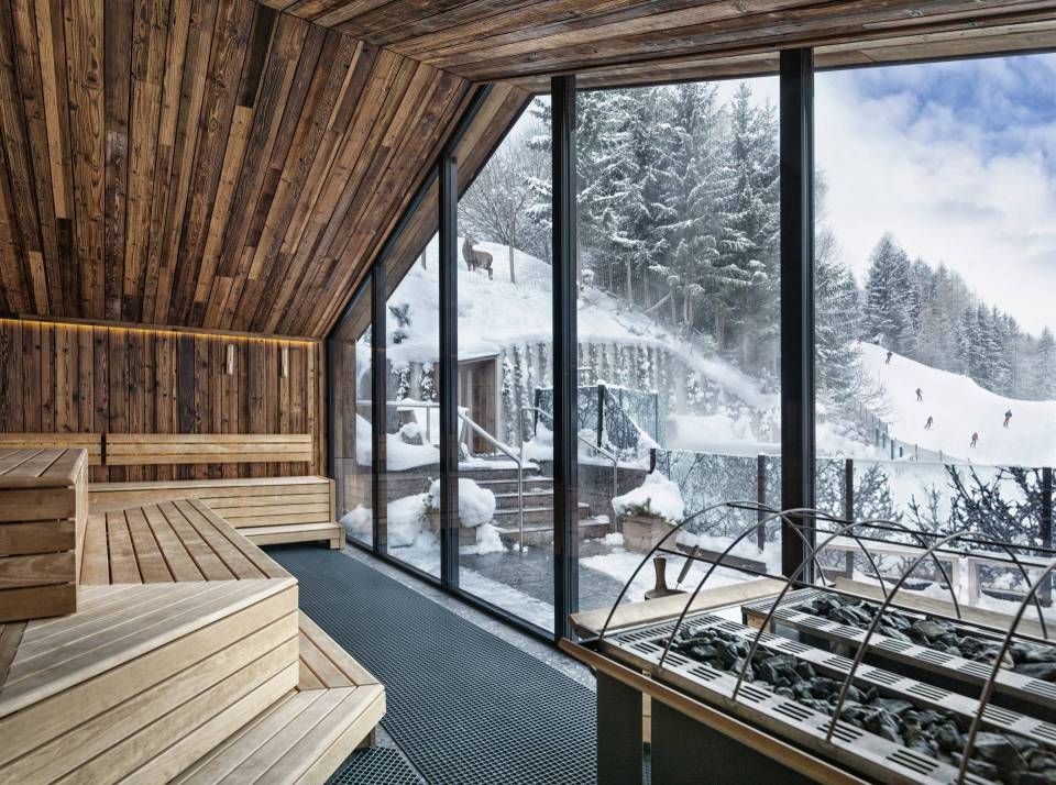 Wooden sauna with large windows overlooking a snowy landscape and skiers.