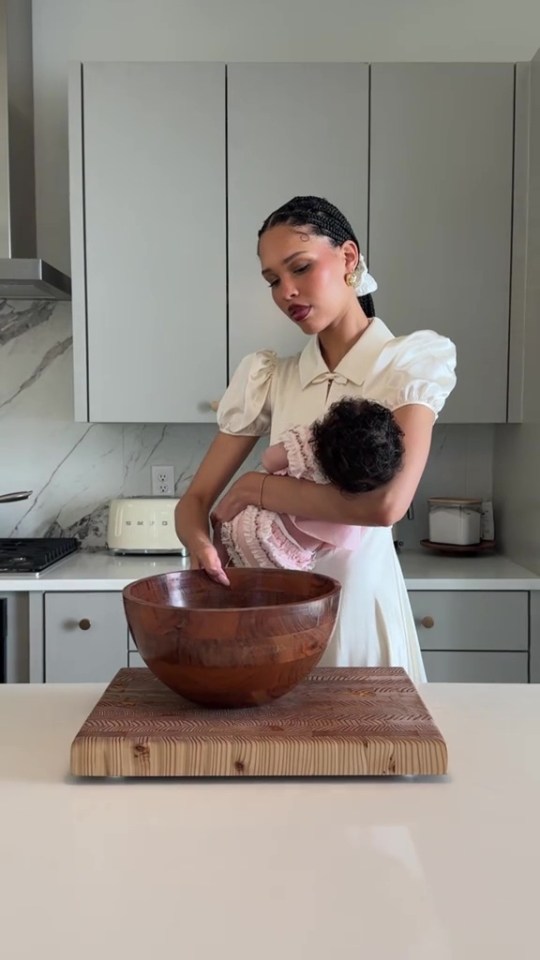 Woman holding baby in kitchen, wooden bowl on cutting board.